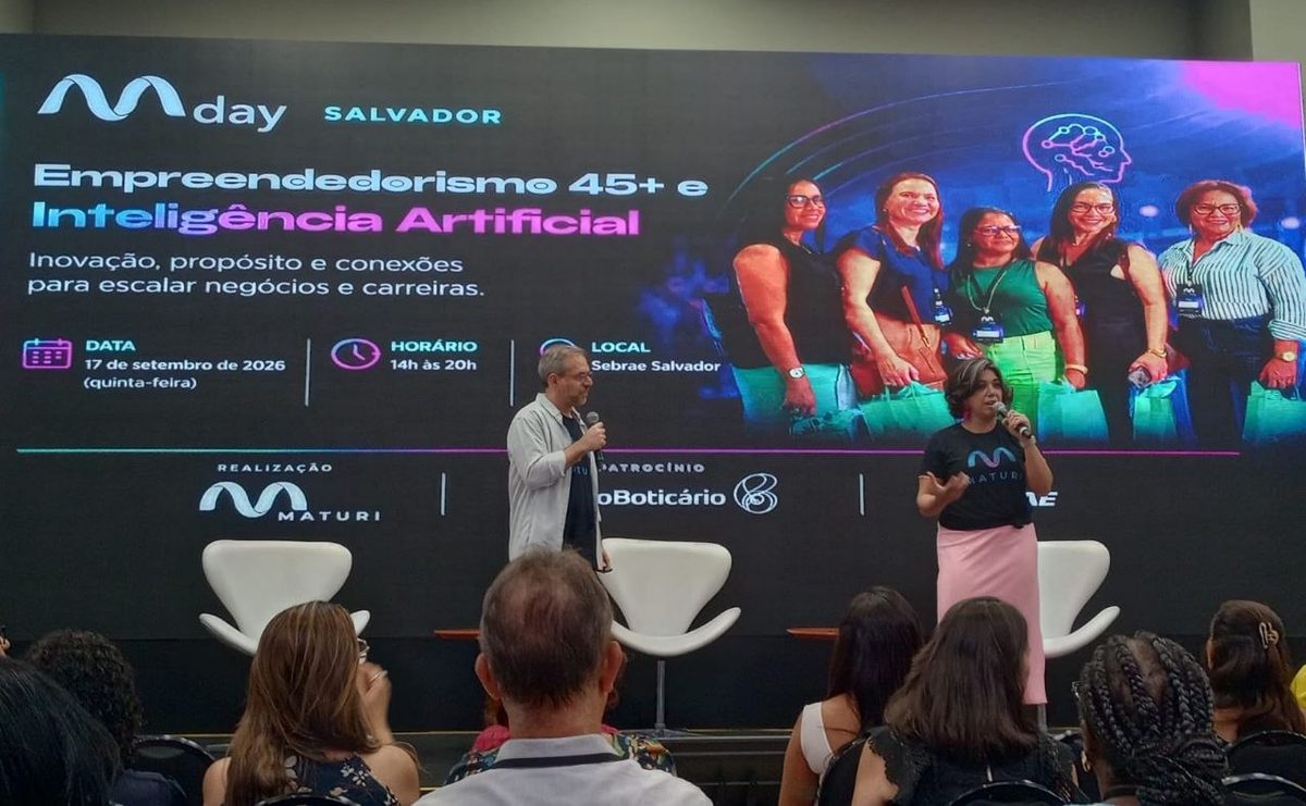 Telão do MaturiDay Salvador anunciando Empreendedorismo 45+ e Inteligência Artificial, com dois apresentadores de microfone em punho diante da plateia no Sebrae Costa Azul